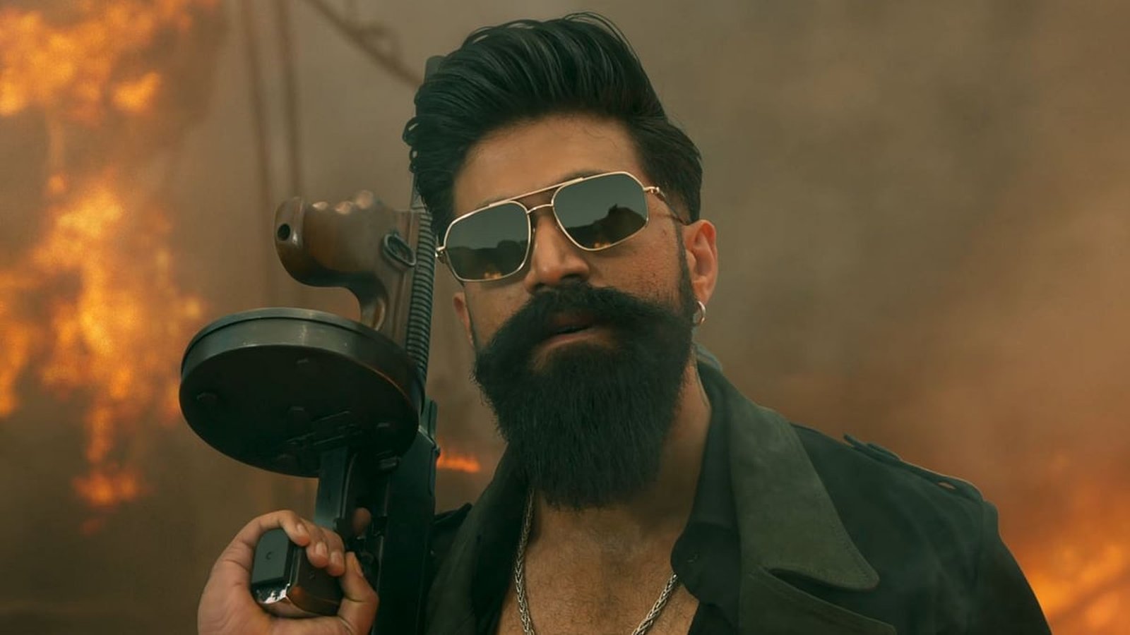 Actor Yash wearing sunglasses and holding a machine gun in a fiery scene