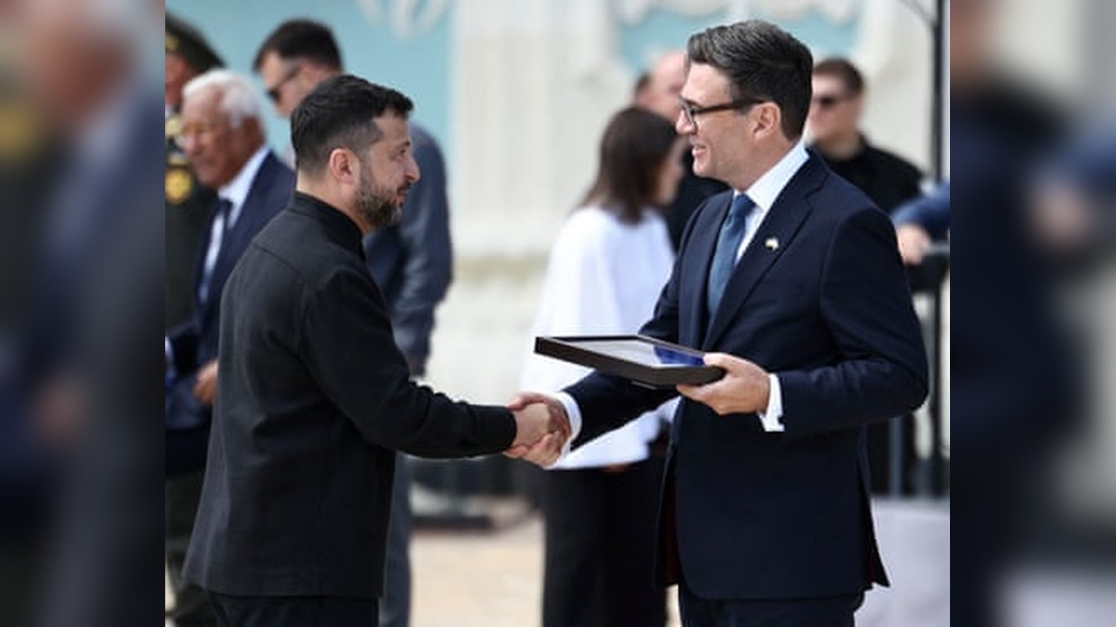 President Volodymyr Zelenskyy shaking hands with a man in a dark suit outdoors