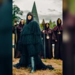 Claudia Winkleman wearing a dark fringed coat standing outdoors with hooded figures holding staffs