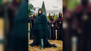 Claudia Winkleman wearing a dark fringed coat standing outdoors with hooded figures holding staffs