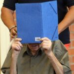 An elderly man, whose face is obscured by a blue folder, is being escorted by another person