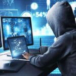 A hooded person sitting at a desk working on multiple computer screens displaying digital security d