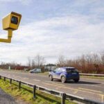 Yellow roadside speed camera mounted on a pole beside a rural Irish road with cars driving past