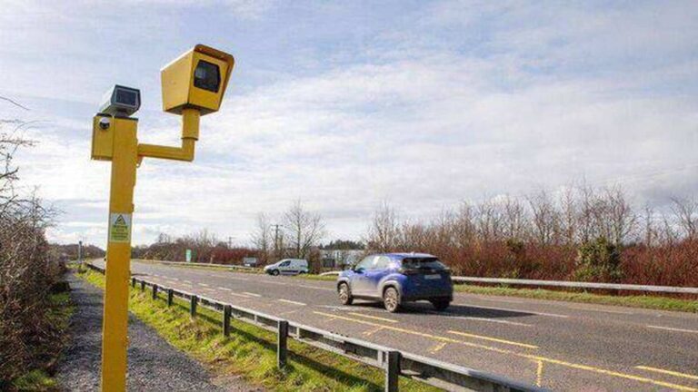 Yellow roadside speed camera mounted on a pole beside a rural Irish road with cars driving past