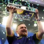 Darts player Luke Littler holding up a trophy with confetti falling around him