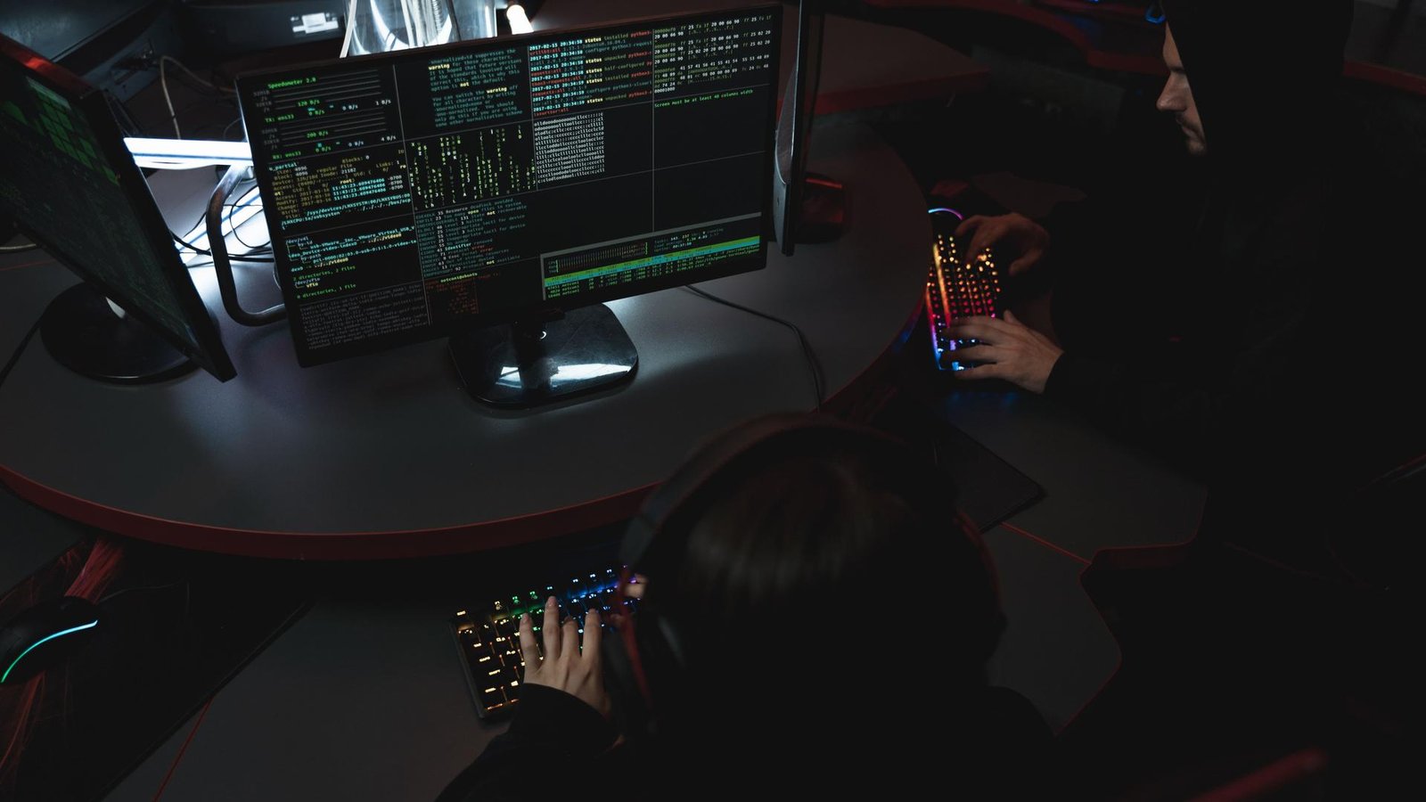 Two hackers working at dual computer monitors displaying lines of green code in a dark room