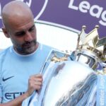 Pep Guardiola holding the Premier League trophy in a light blue Manchester City kit