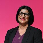 Portrait of Shabana Mahmood smiling against a solid pink backdrop wearing glasses and a dark blazer