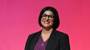 Portrait of Shabana Mahmood smiling against a solid pink backdrop wearing glasses and a dark blazer