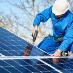 A worker in a blue jumpsuit and white hard hat installs solar panels on a roof using a drill