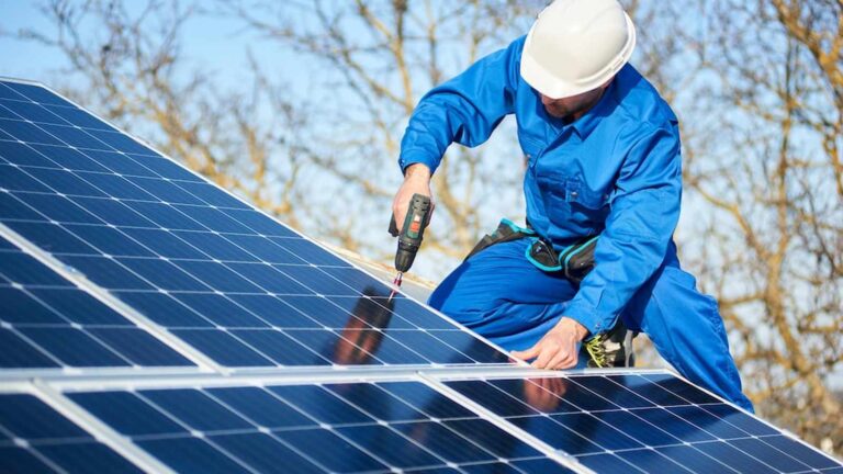 A worker in a blue jumpsuit and white hard hat installs solar panels on a roof using a drill