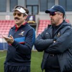 Ted Lasso wearing a visor and sunglasses talking with Coach Beard on a football pitch