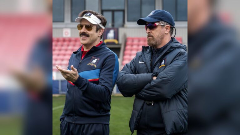 Ted Lasso wearing a visor and sunglasses talking with Coach Beard on a football pitch