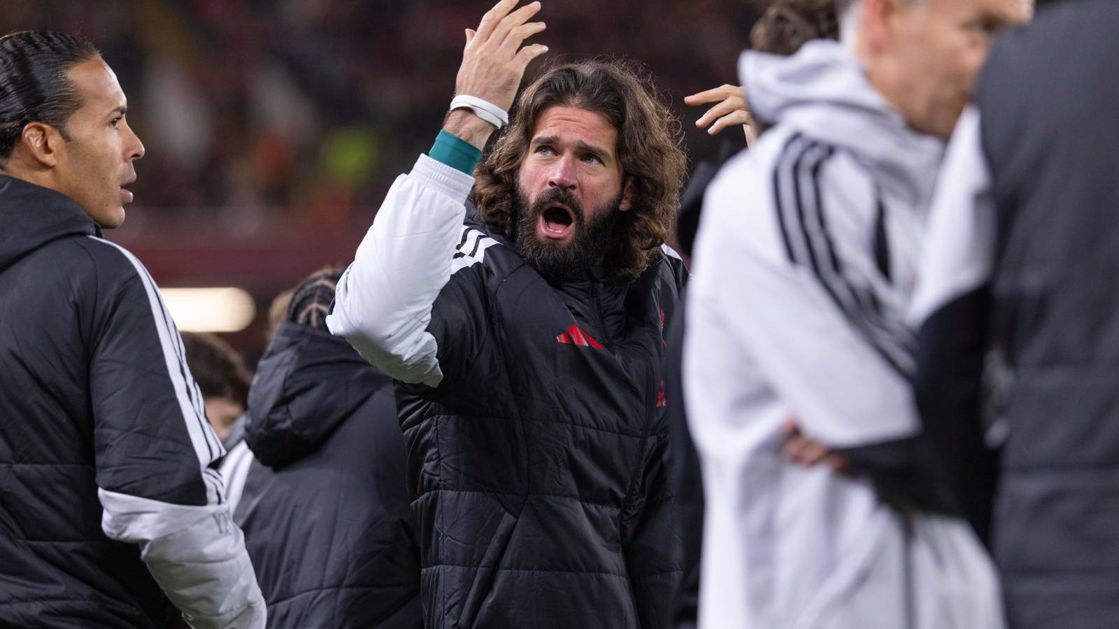 Alisson Becker with long hair and beard gesturing while wearing a black jacket on the pitch