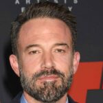 Close up portrait of actor Ben Affleck with a beard in front of a red backdrop