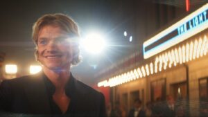A smiling blonde man in a dark suit standing outside a glowing cinema marquee