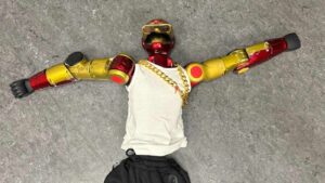 A humanoid robot wearing a white tank top and gold chain lying flat on a grey floor