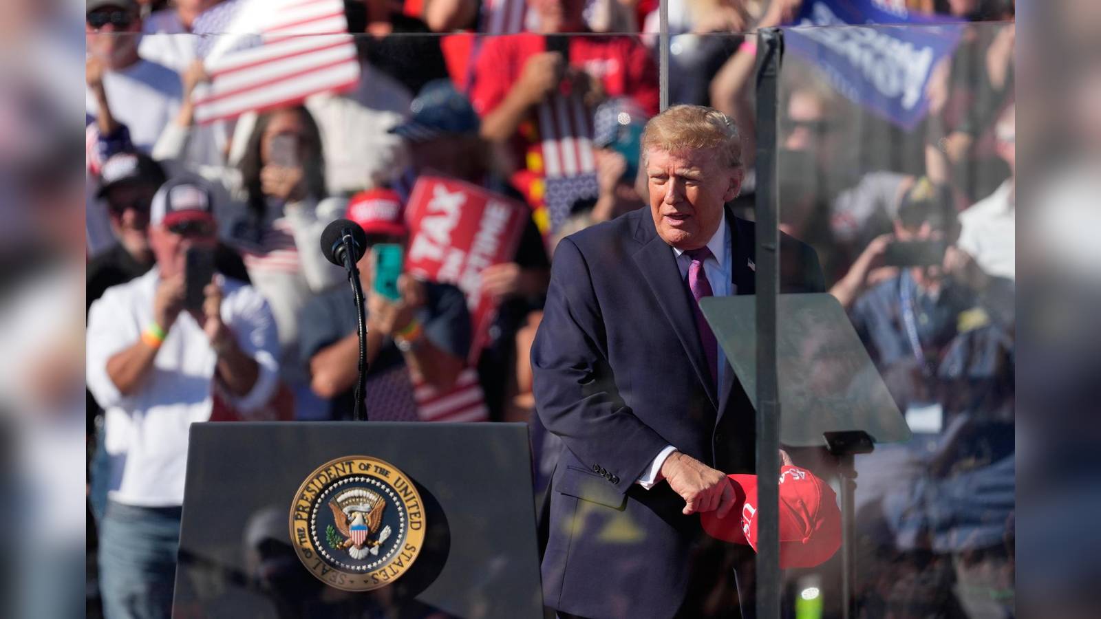 Donald Trump standing behind a podium with presidential seal holding a red hat at a rally with suppo