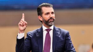 Donald Trump Jr. wearing a blue suit and purple tie raises a finger while speaking