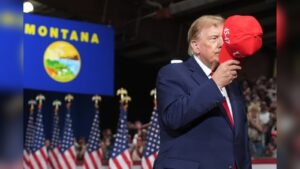 Donald Trump wearing a blue suit holds a red hat at a campaign rally in front of a Montana flag