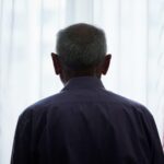 An elderly person viewed from behind looking out a bright window through sheer curtains