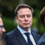 Close-up of Elon Musk wearing a dark suit and blue tie surrounded by people