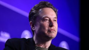 Elon Musk speaking at an event against a blue backdrop