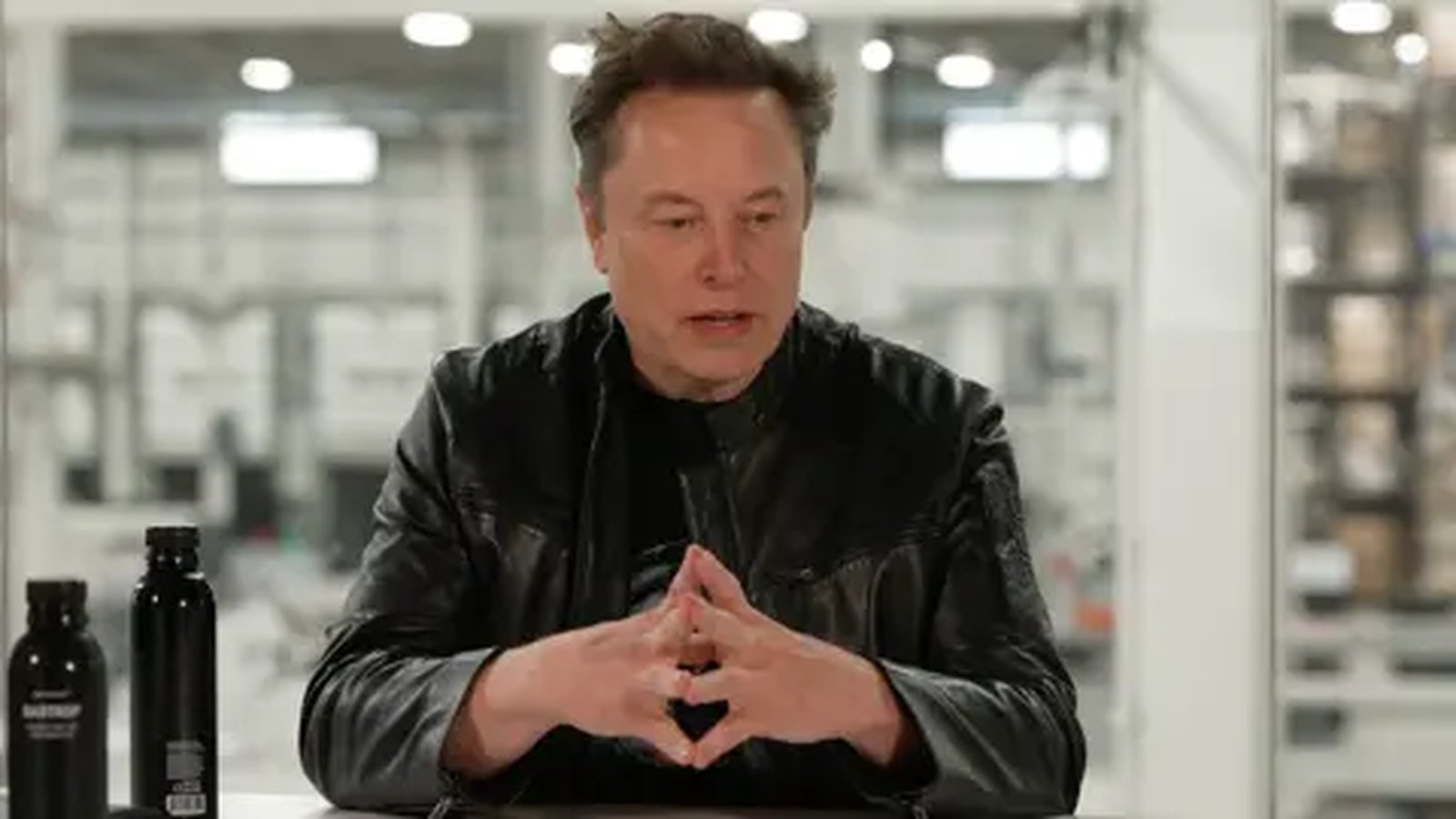 Elon Musk sitting indoors wearing a black jacket with hands clasped