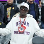 Rapper Flavor Flav wearing sunglasses and a white hoodie reacting at a basketball game