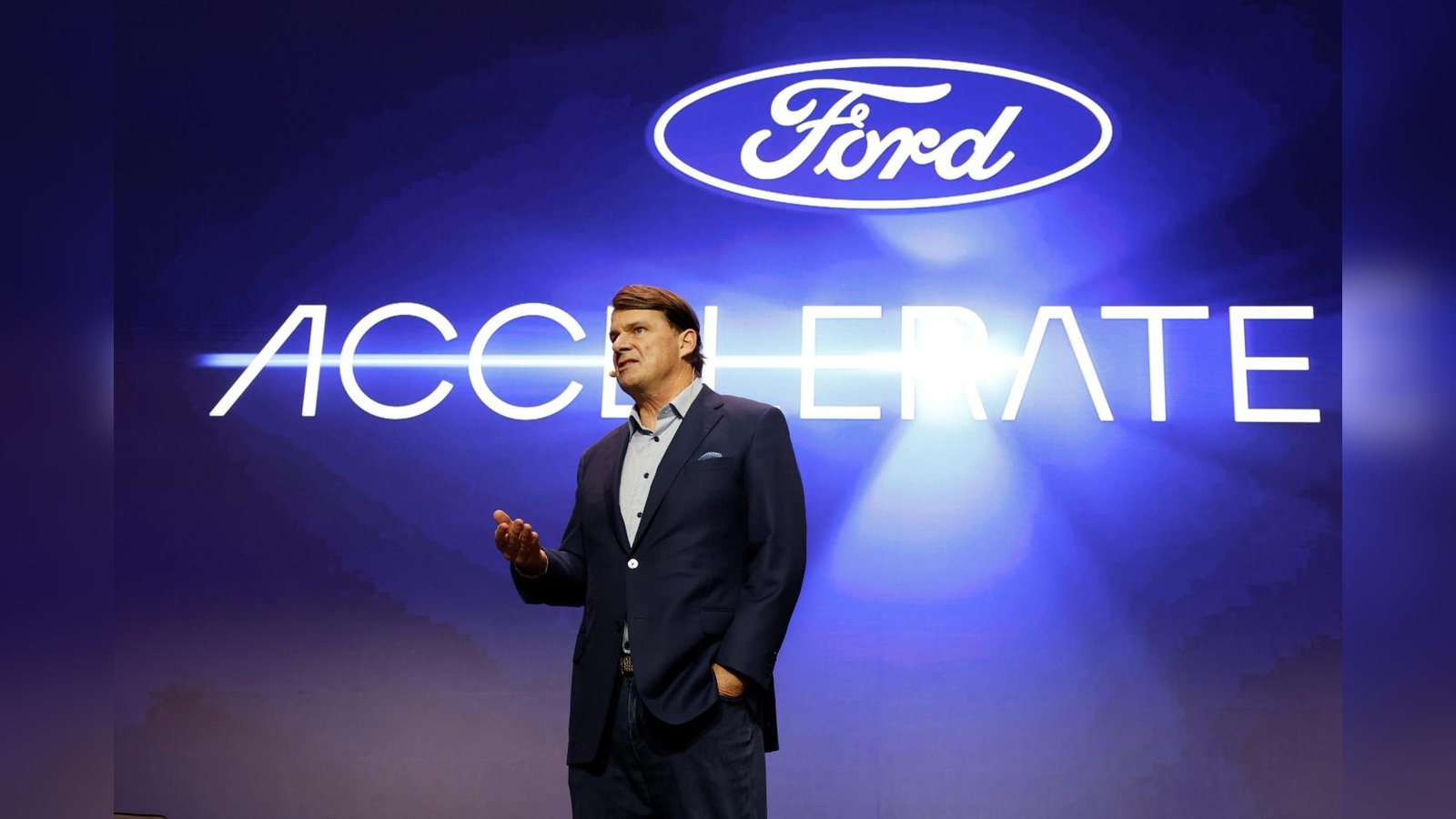 Ford CEO Jim Farley speaking on stage in front of a blue corporate background