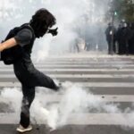 A protester in black clothing kicks away tear gas amidst smoke with police officers standing in the