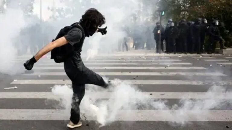A protester in black clothing kicks away tear gas amidst smoke with police officers standing in the