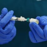 A healthcare worker wearing blue medical gloves prepares a vaccine injection from a small vial