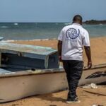 An IOM worker in a branded t-shirt inspecting a wrecked migrant boat on a beach