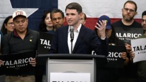 Texas Senate candidate James Talarico speaking at a podium with campaign supporters holding signs