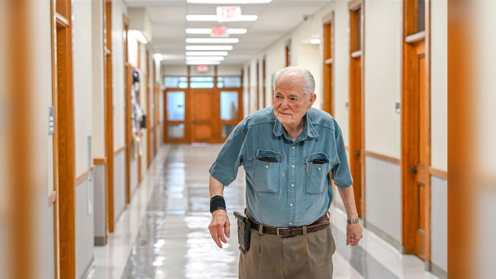 An elderly man in a blue button-down shirt walking down a wood-trimmed university hallway