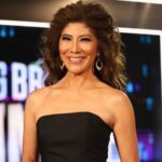 Julie Chen Moonves smiling on the Big Brother set in front of a lit backdrop