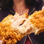 Close-up of hands pulling apart a crispy fried chicken tender showing juicy white meat inside