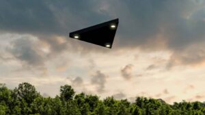 A mysterious matte black triangular craft with glowing lights hovers in a cloudy sky above a forest