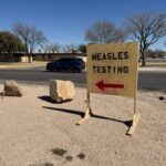 A wooden sign reading Measles Testing with a red arrow pointing left outdoors