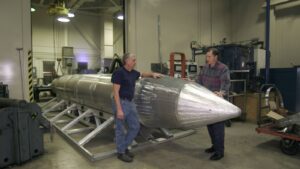 Two men standing next to a large GBU-43B MOAB bomb casing inside a manufacturing workshop