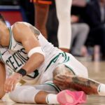 A professional basketball player grimacing in pain while sitting on the hardwood court floor