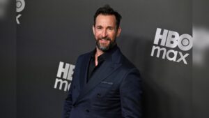 Actor Noah Wyle smiling in a dark suit jacket in front of an HBO Max backdrop