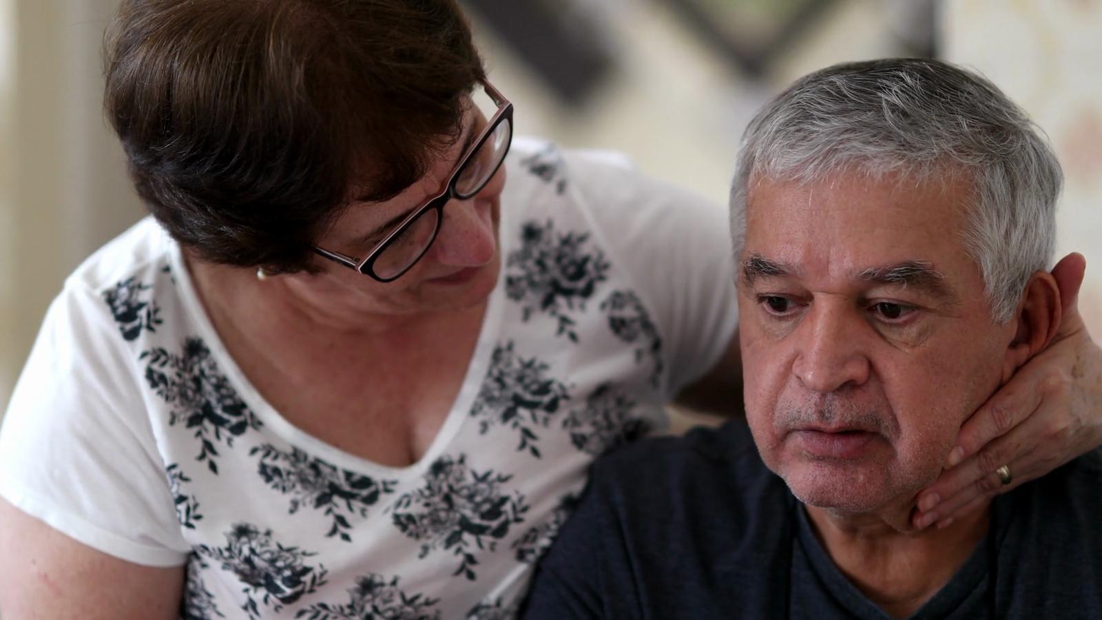 An older woman supporting her concerned older husband with her hand on his neck