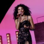 Singer Olivia Dean performing live on stage with a pink background and microphone