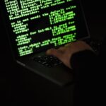 A person typing on a laptop keyboard with green code displayed on the screen in a dark room