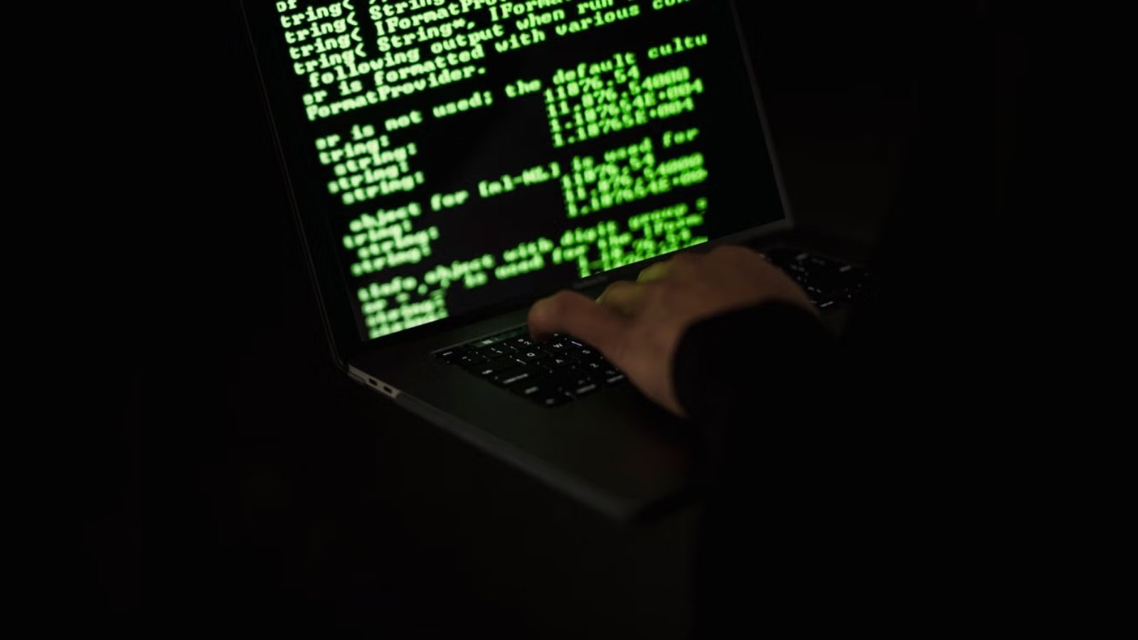 A person typing on a laptop keyboard with green code displayed on the screen in a dark room