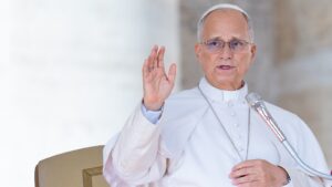 Pope Leo XIV wearing white vestments and glasses speaking into a microphone while raising his hand