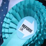 Close up of a turquoise Reform UK rosette pinned to a dark suit jacket