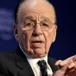 Rupert Murdoch wearing glasses and a suit speaking at a conference with a microphone